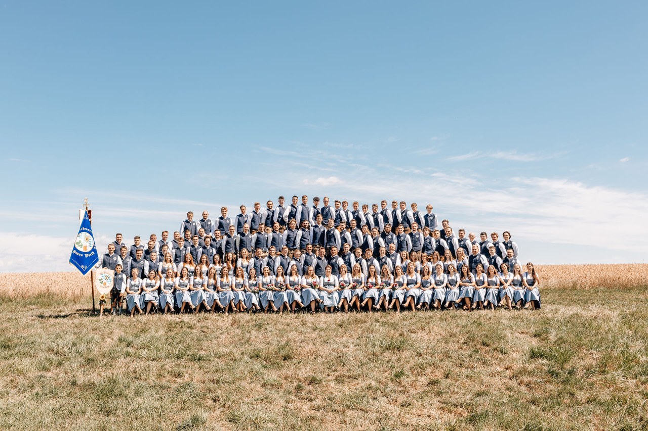 Gruppenbild des Vereins mit Festdamen und Vereinsfahne vor blauem Himmel